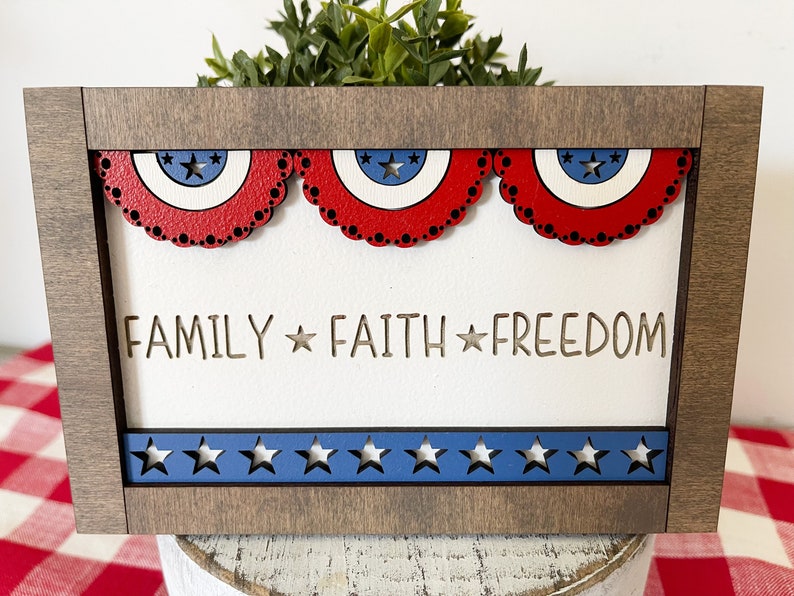 May include: A wooden framed sign with a white background and the words "FAMILY * FAITH * FREEDOM" in a cutout font. The sign is decorated with red, white, and blue stars and banners.