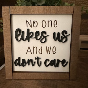 Puede incluir: Un letrero de madera con el texto "No one likes us and we don't care" en letras negras sobre fondo blanco.