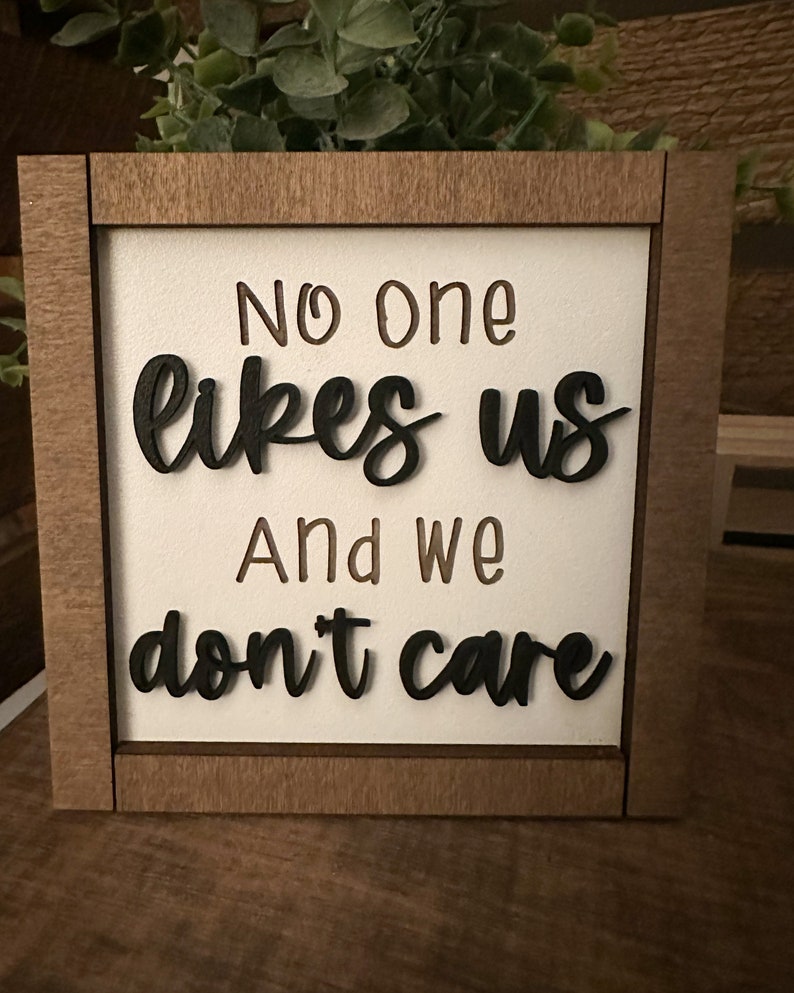 Puede incluir: Un letrero de madera con el texto "No one likes us and we don't care" en letras negras sobre fondo blanco.