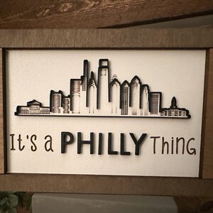 Puede incluir: Un letrero de madera con una silueta negra del horizonte de Filadelfia y el texto "It's a Philly Thing".