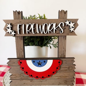 May include: Wooden sign with the words "FIREWORKS" in white letters. The sign is decorated with red, white, and blue stars and a red, white, and blue bunting.