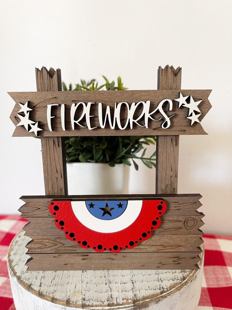 May include: Wooden sign with the words "FIREWORKS" in white letters. The sign is decorated with red, white, and blue stars and a red, white, and blue bunting.