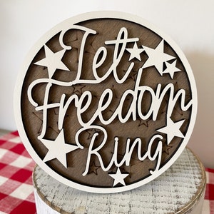 May include: A white wooden round sign with the text "Let Freedom Ring" in white lettering. The sign is decorated with white stars.