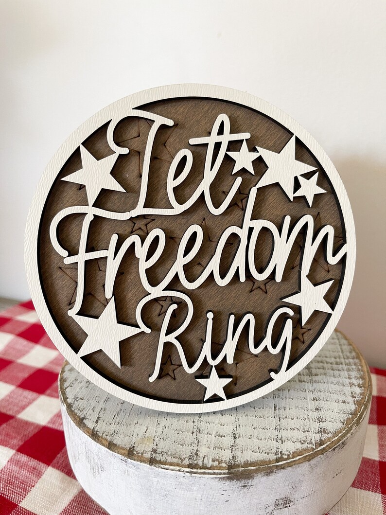 May include: A white wooden round sign with the text "Let Freedom Ring" in white lettering. The sign is decorated with white stars.