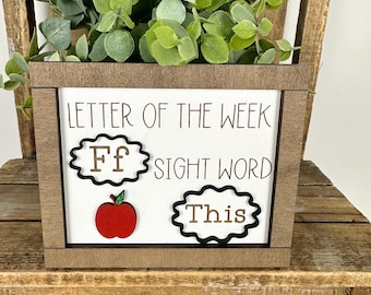 May include: A wooden box with the text "LETTER OF THE WEEK" and "SIGHT WORD" on a white background. The box features the letter "Ff", a red apple, and the word "This". Green foliage sits atop the box.
