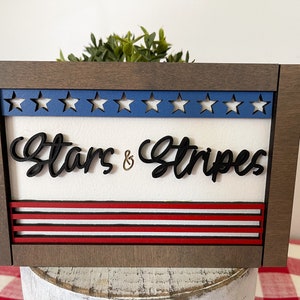 May include: A wooden framed sign with a white background and the words "Stars & Stripes" in black lettering. The top of the sign has a blue and white star design and the bottom has red and white stripes.