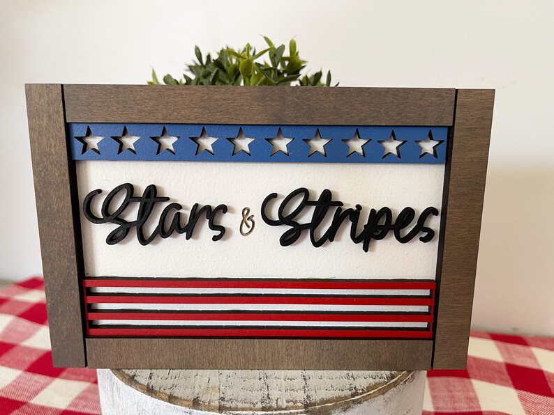 May include: A wooden framed sign with a white background and the words "Stars & Stripes" in black lettering. The top of the sign has a blue and white star design and the bottom has red and white stripes.