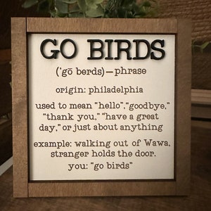 Puede incluir: Un letrero de madera con el texto "GO BIRDS" en letras negras. El letrero define la frase "Go Birds" como un t&eacute;rmino de jerga de Filadelfia que se puede usar para decir "hola", "adi&oacute;s", "gracias", "que tengas un buen d&iacute;a", o casi cualquier cosa. El letrero incluye un ejemplo de la frase utilizada en una conversaci&oacute;n.
