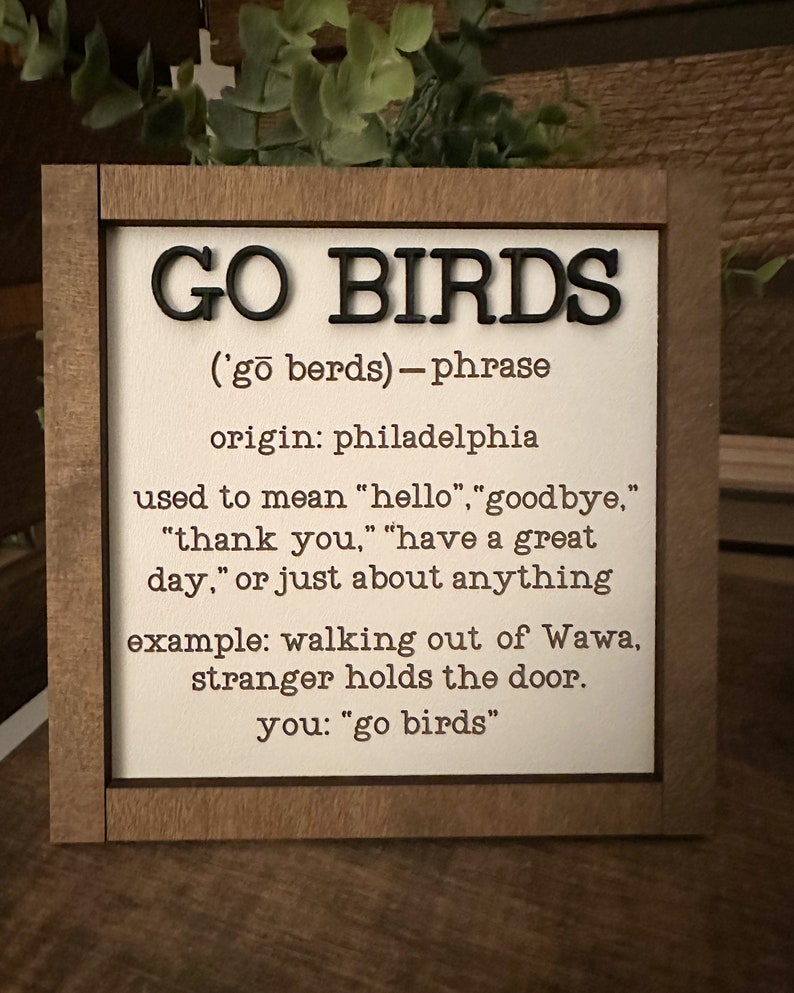 Puede incluir: Un letrero de madera con el texto "GO BIRDS" en letras negras. El letrero define la frase "Go Birds" como un t&eacute;rmino de jerga de Filadelfia que se puede usar para decir "hola", "adi&oacute;s", "gracias", "que tengas un buen d&iacute;a", o casi cualquier cosa. El letrero incluye un ejemplo de la frase utilizada en una conversaci&oacute;n.