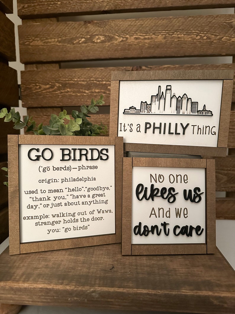 Puede incluir: Tres letreros de madera con letras negras. El primer letrero dice "GO BIRDS ('go berds) - phrase origin: philadelphia used to mean "hello", "goodbye", "thank you", "have a great day", or just about anything example: walking out of Wawa, stranger holds the door. you: "go birds"". El segundo letrero dice "It's a PHILLY Thing". El tercer letrero dice "No one likes us And we don't care".