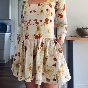 May include: A cream-colored dress with long sleeves and a square neckline. The dress is adorned with a floral pattern in shades of brown, orange, and green. The skirt has a flared, ruffled design. The dress has pockets.