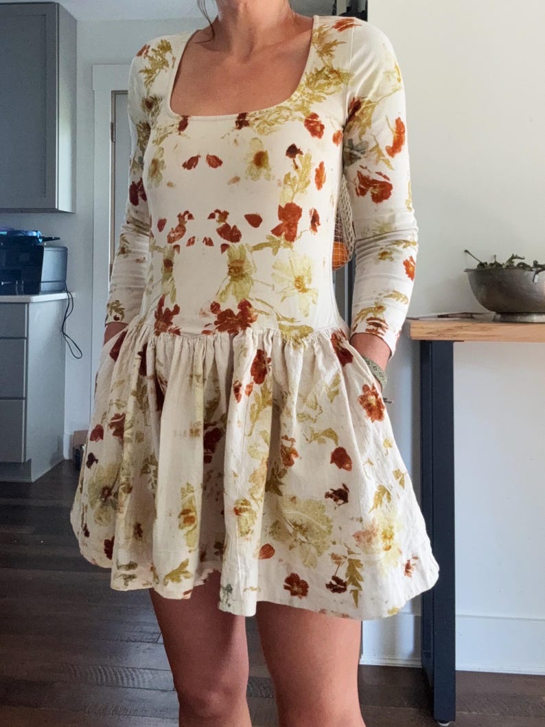 May include: A cream-colored dress with long sleeves and a square neckline. The dress is adorned with a floral pattern in shades of brown, orange, and green. The skirt has a flared, ruffled design. The dress has pockets.