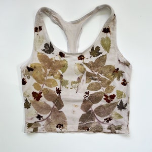 May include: A beige racerback crop top featuring a botanical print of various leaves and small dark red flowers. The garment has a scoop neckline and a sleeveless design, with the natural plant-based patterns distributed across the front surface.