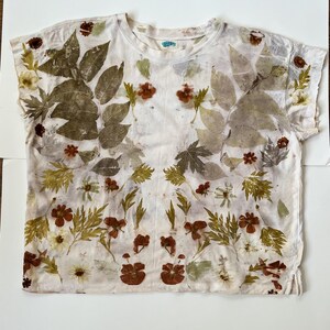 May include: A short-sleeved t-shirt in an off-white colour featuring a botanical print of various pressed leaves and flowers in shades of olive green, brown, and rust orange. The garment has a crew neckline and a straight hem with side slits.