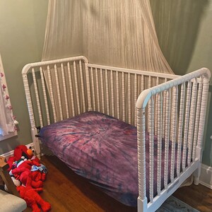 May include: White wooden crib with a purple and pink tie-dye sheet. The crib has a white canopy hanging over it.