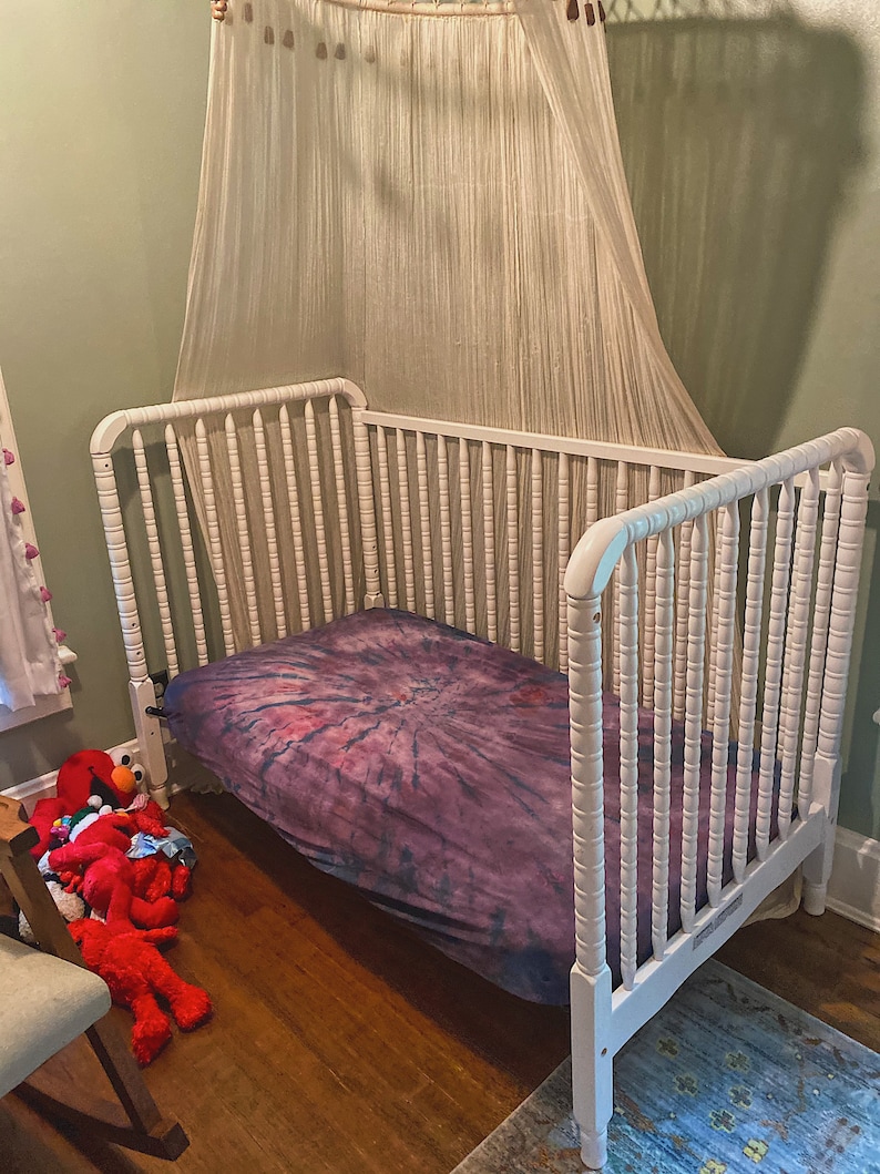 May include: White wooden crib with a purple and pink tie-dye sheet. The crib has a white canopy hanging over it.