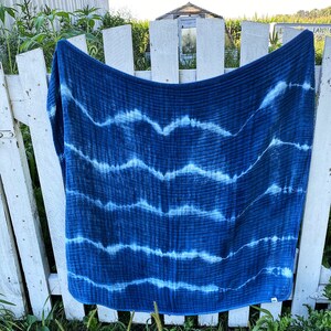 May include: A blue and white tie-dye cotton baby blanket hanging on a white picket fence. The blanket has a wavy pattern and is made of soft, breathable fabric.