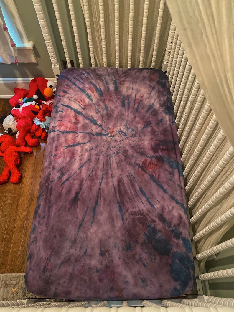 May include: A purple and blue tie-dye fitted crib sheet with a swirling pattern.
