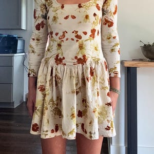 May include: A long-sleeved, cream-colored dress with a square neckline and a gathered skirt. The dress is adorned with a floral pattern in shades of red, brown, and yellow. The dress is knee-length.