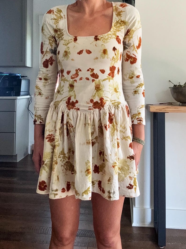 May include: A long-sleeved, cream-colored dress with a square neckline and a gathered skirt. The dress is adorned with a floral pattern in shades of red, brown, and yellow. The dress is knee-length.