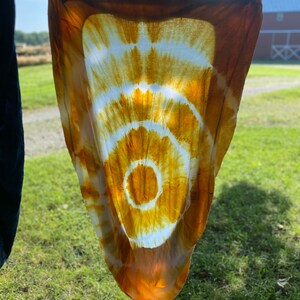 May include: A white fabric with a large, circular tie-dye pattern in shades of orange and yellow. The fabric is hanging on a clothesline outdoors.