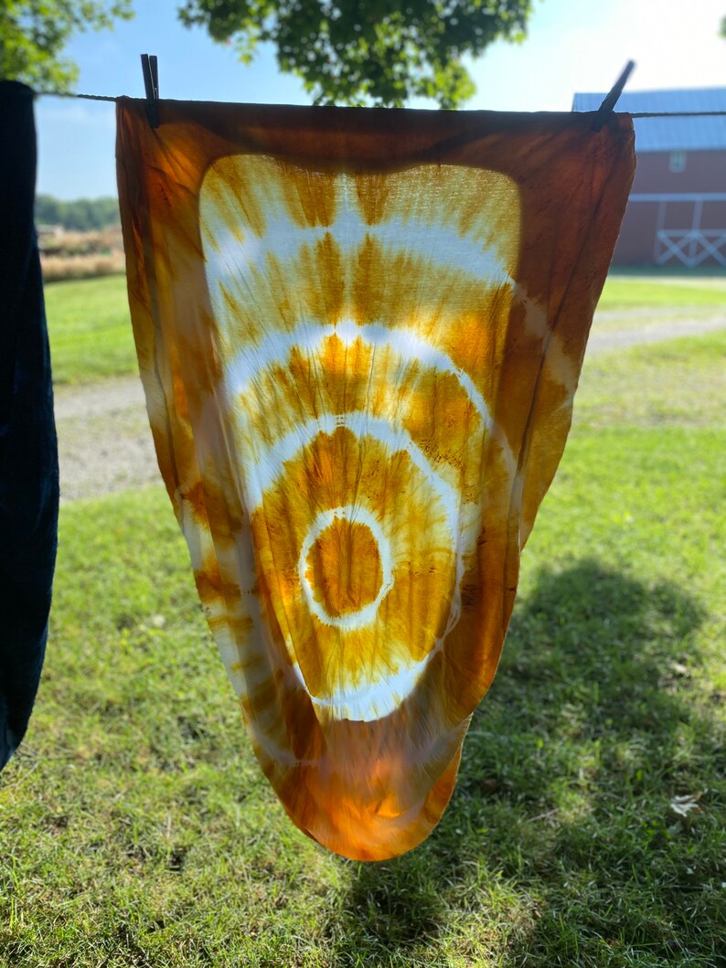 May include: A white fabric with a large, circular tie-dye pattern in shades of orange and yellow. The fabric is hanging on a clothesline outdoors.