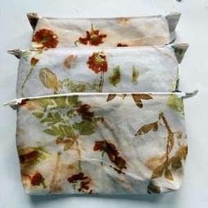 May include: Three canvas zipper pouches with botanical prints. The pouches feature a cream-colored background with pressed flowers and leaves in shades of orange, brown, and green. Each pouch has a white zipper and a rectangular shape.