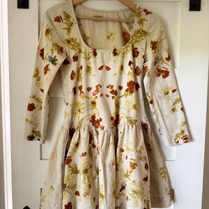May include: A cream-colored dress with long sleeves and a square neckline, adorned with a floral pattern in shades of red, green, and yellow. The dress features a ruffled skirt and is hanging on a wooden hanger.
