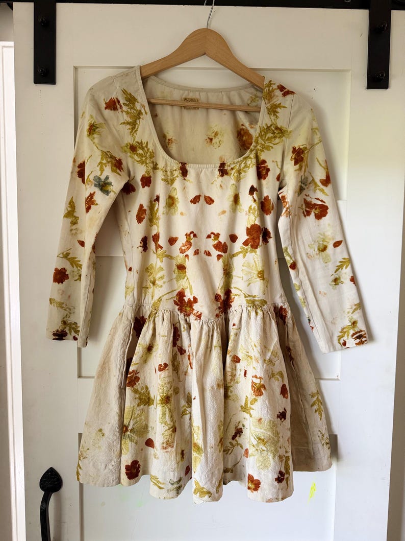 May include: A cream-colored dress with long sleeves and a square neckline, adorned with a floral pattern in shades of red, green, and yellow. The dress features a ruffled skirt and is hanging on a wooden hanger.