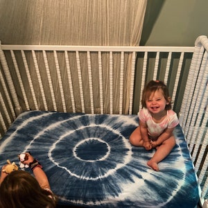 May include: A white crib with a blue and white tie-dye crib sheet. A young child is sitting on the sheet smiling.