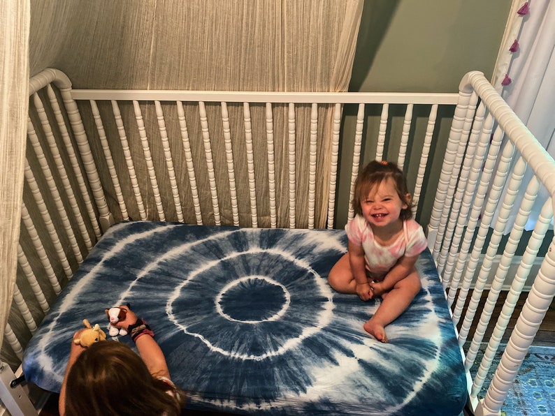 May include: A white crib with a blue and white tie-dye crib sheet. A young child is sitting on the sheet smiling.