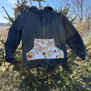 May include: A dark green hooded jacket with a zipper closure and a large pocket on the front. The pocket is lined with a white fabric with a floral pattern.
