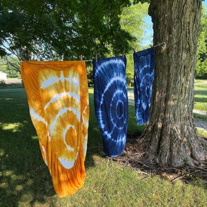 May include: Three tie-dye fabric panels hanging on a clothesline outside. The panels are in shades of orange, blue, and white.