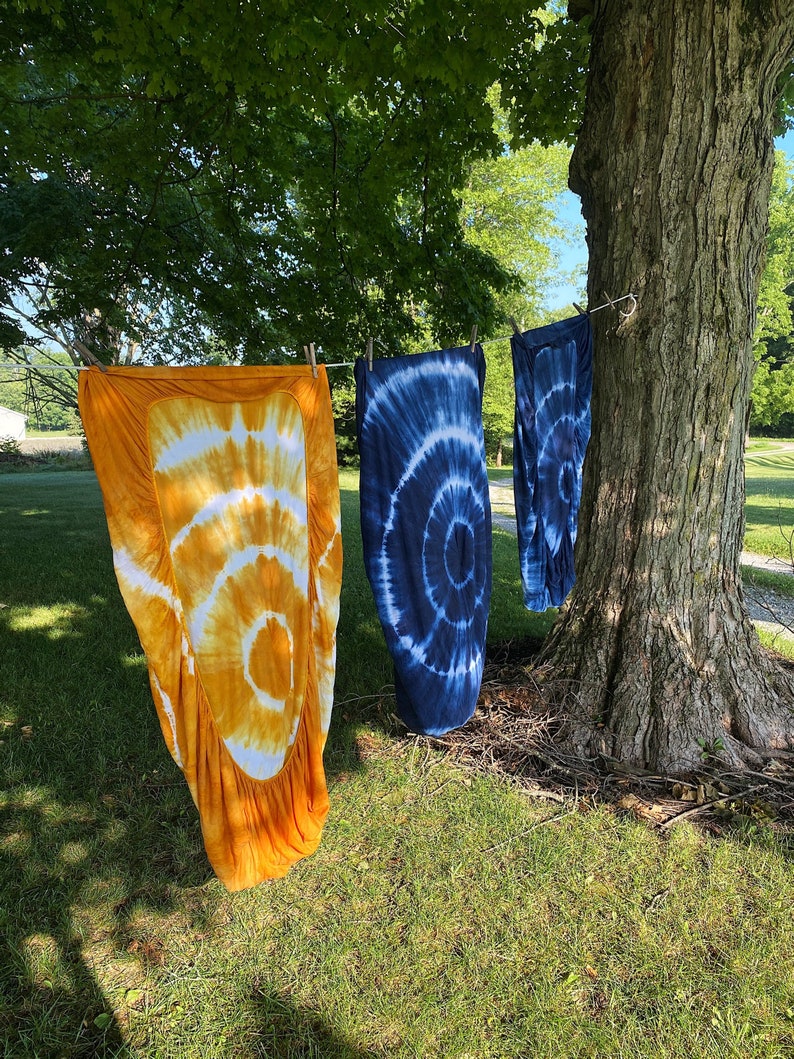 May include: Three tie-dye fabric panels hanging on a clothesline outside. The panels are in shades of orange, blue, and white.