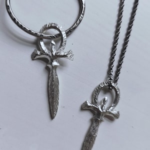 May include: A silver necklace and earrings set featuring an ankh pendant with a textured finish. The necklace has a silver chain and the earrings are hoop style.