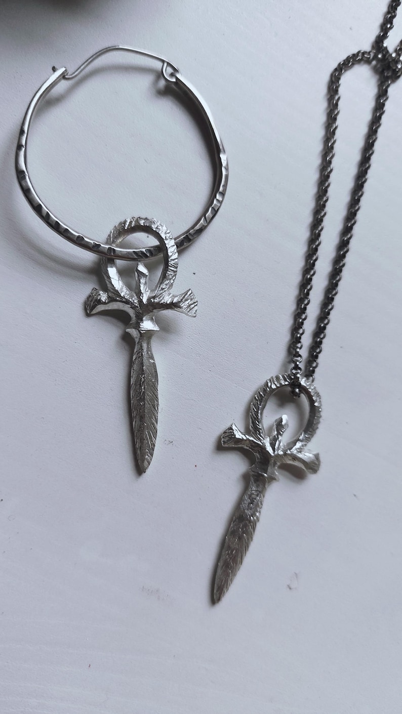 May include: A silver necklace and earrings set featuring an ankh pendant with a textured finish. The necklace has a silver chain and the earrings are hoop style.