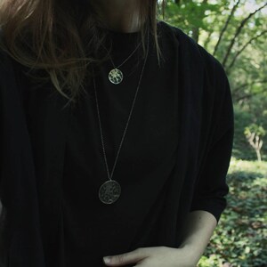 May include: A person wearing a black shirt with two silver necklaces. The necklaces have round, textured pendants.
