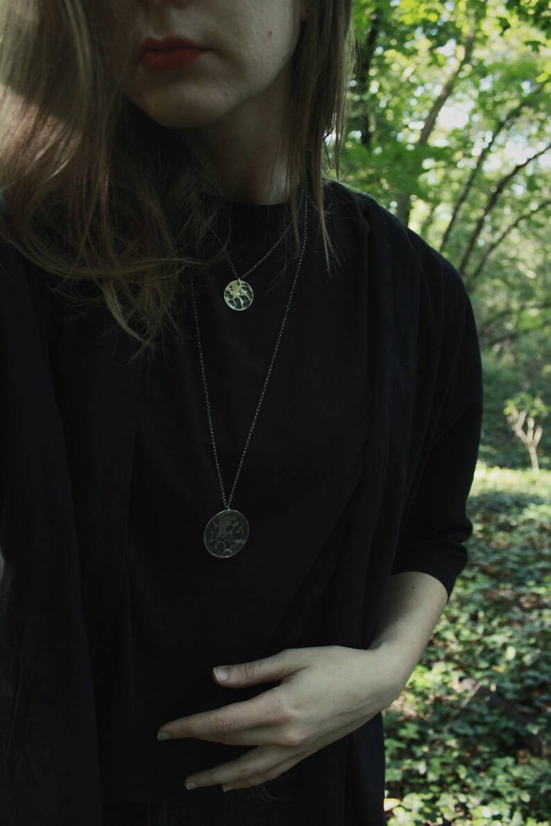 May include: A person wearing a black shirt with two silver necklaces. The necklaces have round, textured pendants.
