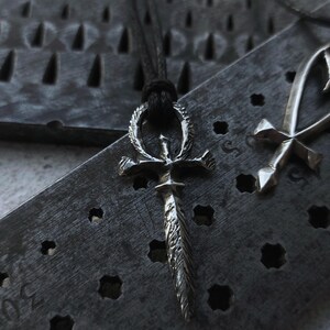 May include: A silver Ankh pendant with a detailed design, hanging on a black cord. The pendant is resting on a textured metal surface.