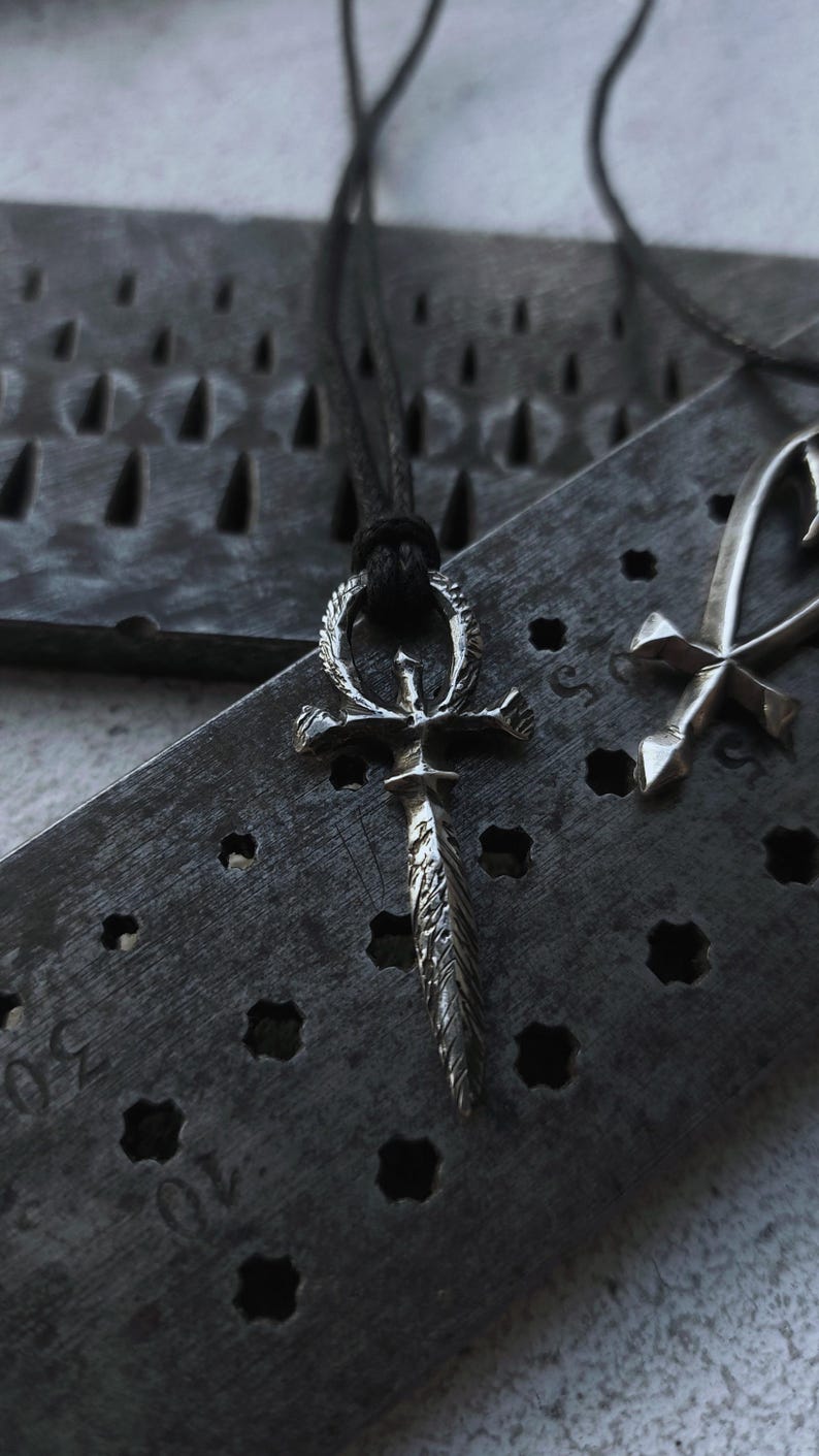 May include: A silver Ankh pendant with a detailed design, hanging on a black cord. The pendant is resting on a textured metal surface.
