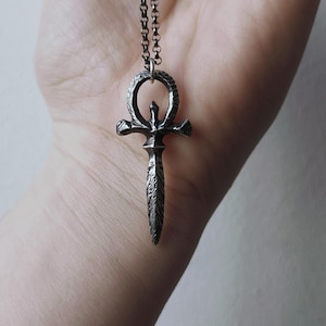 May include: A silver chain necklace with a pendant featuring a stylized ankh symbol with a dagger-like point.