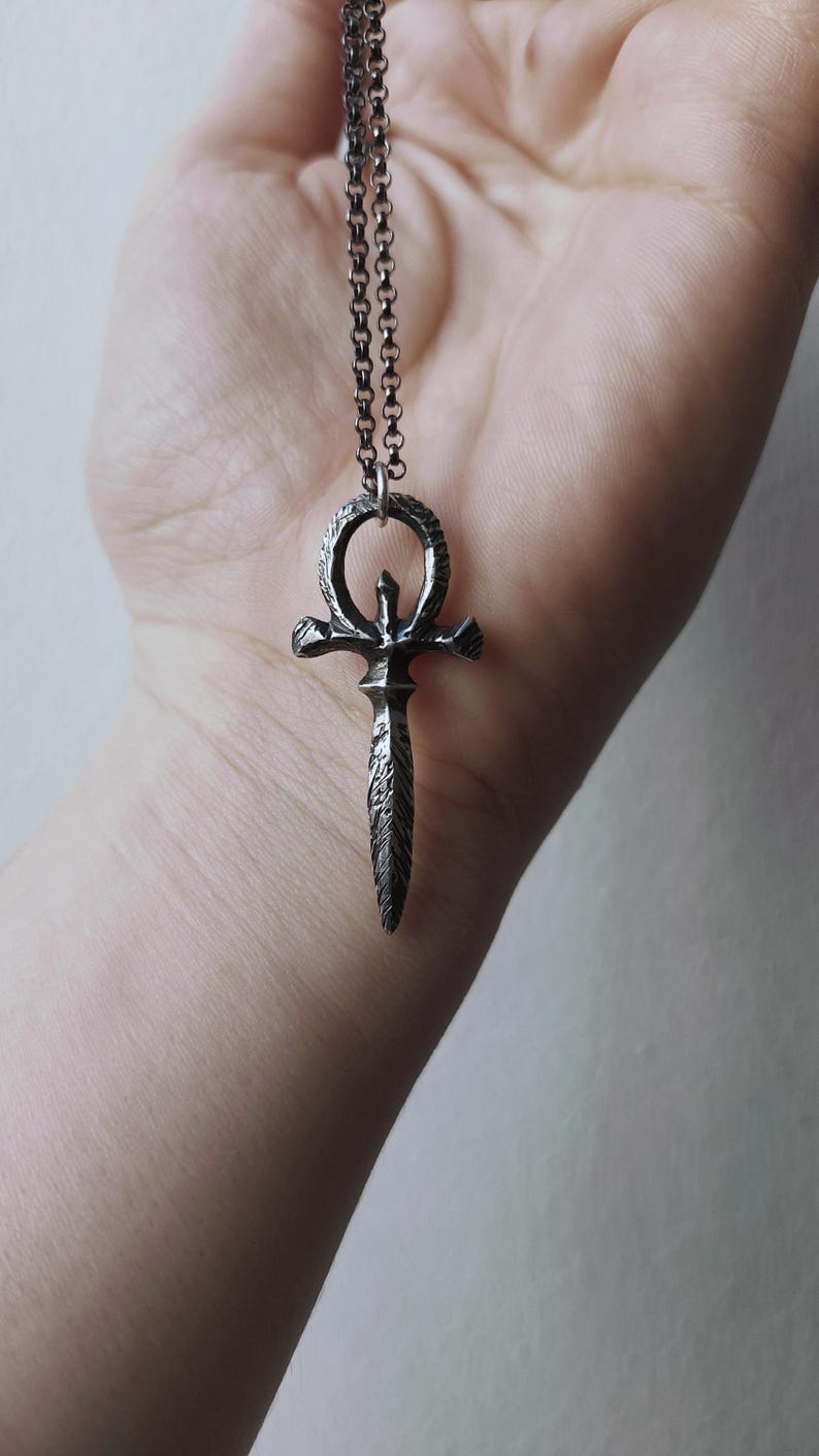 May include: A silver chain necklace with a pendant featuring a stylized ankh symbol with a dagger-like point.