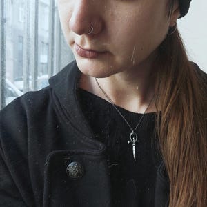 May include: A person wearing a black jacket with a white stripe and a silver necklace with a dagger pendant. The person has a nose ring and a black beanie.
