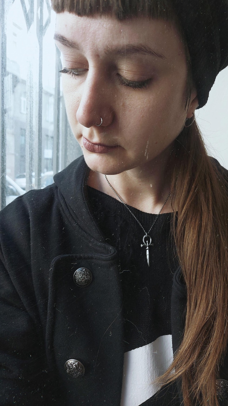 May include: A person wearing a black jacket with a white stripe and a silver necklace with a dagger pendant. The person has a nose ring and a black beanie.