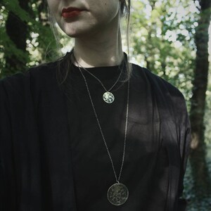 May include: A person wearing a black top with two silver necklaces. The necklaces have round, textured pendants.