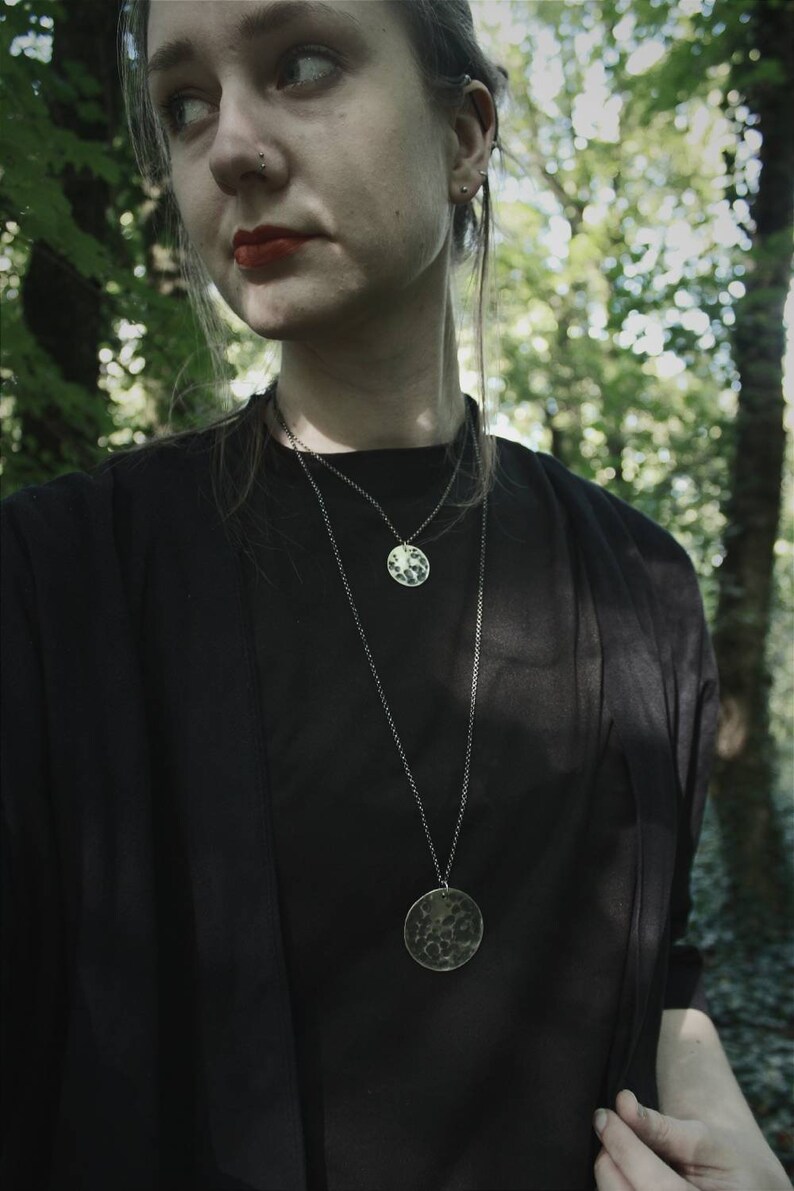 May include: A person wearing a black top with two silver necklaces. The necklaces have round, textured pendants.
