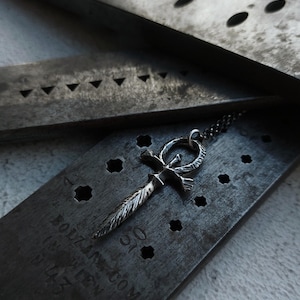 May include: A silver ankh pendant with a feather design hangs from a silver chain. The pendant is resting on a metal surface with holes and the text "HOLZANDTOM.COM" and "MADE WITH LOVE" is visible.