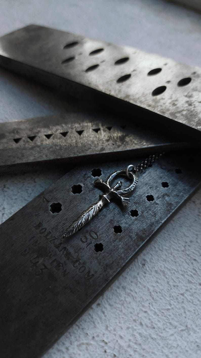 May include: A silver ankh pendant with a feather design hangs from a silver chain. The pendant is resting on a metal surface with holes and the text "HOLZANDTOM.COM" and "MADE WITH LOVE" is visible.