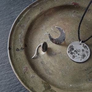 May include: Three silver jewelry pieces on a green metal plate. The pieces include a crescent moon earring, a small oval earring, and a round pendant with a textured surface.