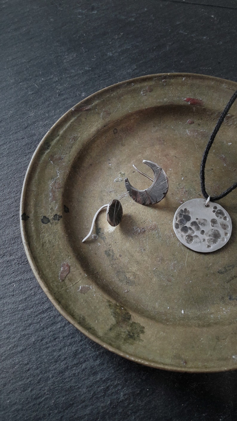 May include: Three silver jewelry pieces on a green metal plate. The pieces include a crescent moon earring, a small oval earring, and a round pendant with a textured surface.